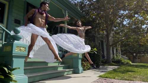 Two Blue13 dancers, wearing white hoop skirts, jump from the steps of a building at Heritage Square museum, reaching arms toward each other while in midair.