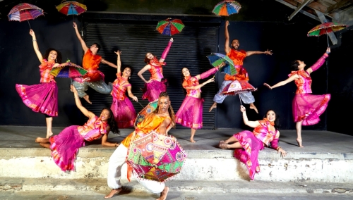 Blue13 dancers pose for a shot, dressed in pink and orange traditional Indian garb, with several holding colorful parasols